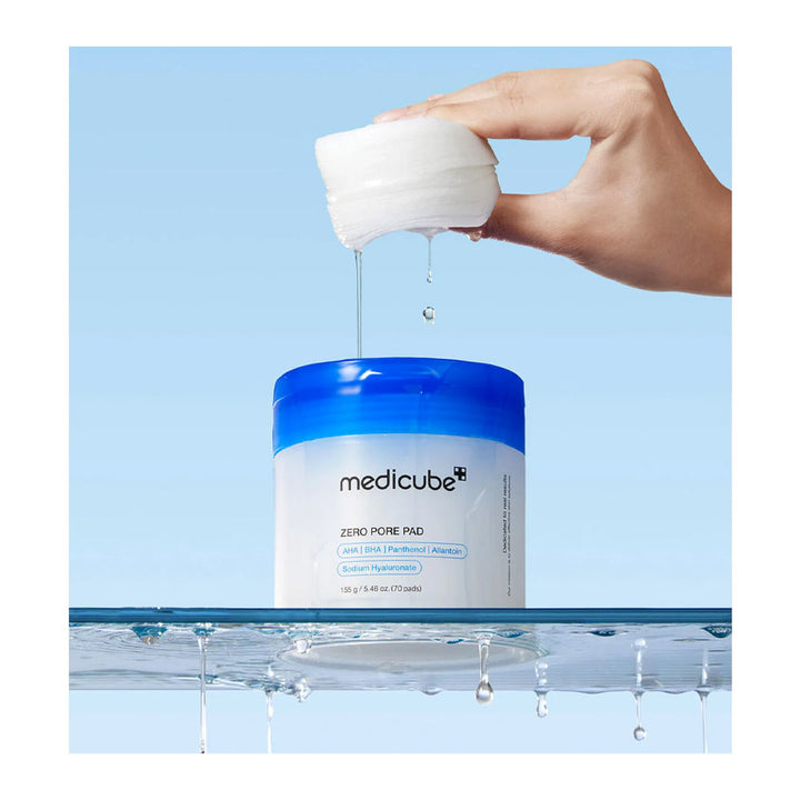 A hand holds a white pad above a blue-and-white medicube Zero Pore Pad jar, with a drop of clear gel dripping between them, set against a light blue background with water droplets at the bottom.