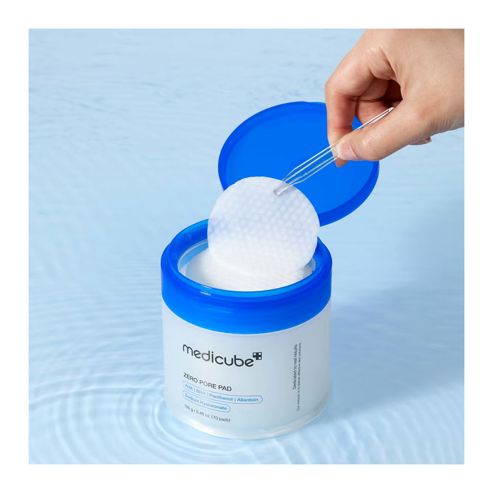 A hand uses tweezers to lift a round white cleansing pad from an open blue-and-white Medicube Zero Pore Pad container, set against a light blue water-textured background.