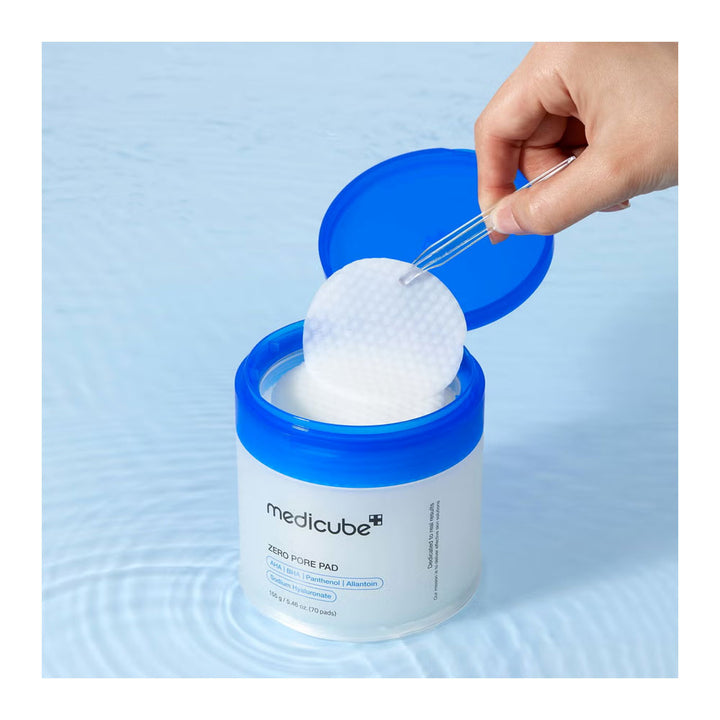 A hand uses tweezers to lift a round white cleansing pad from an open blue-and-white Medicube Zero Pore Pad container, set against a light blue water-textured background.