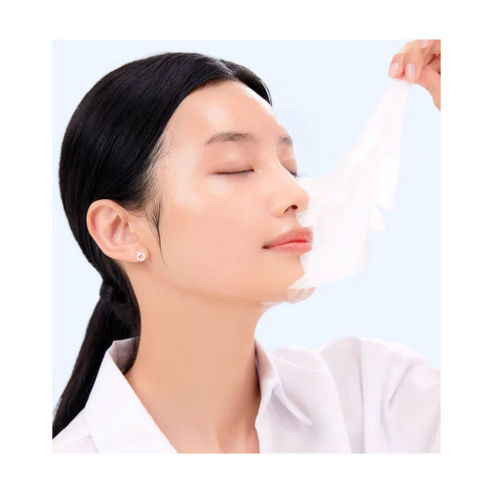 A woman with closed eyes and a white shirt peels a translucent, skin-like sheet from her cheek against a light blue background.