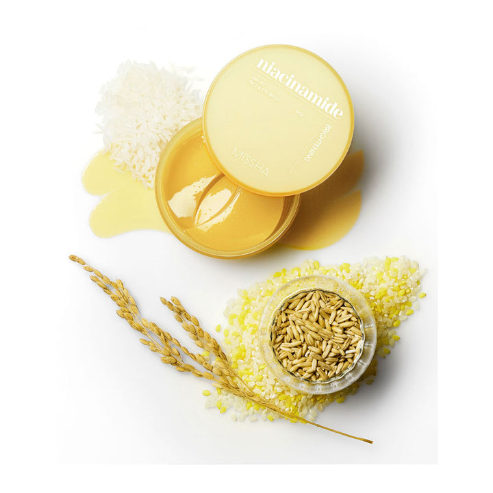 A round yellow cosmetic jar with its lid off, showing a golden-yellow cream inside, styled on a white background with oats, oat flakes, and wheat stalks. The lid is printed with the brand name &