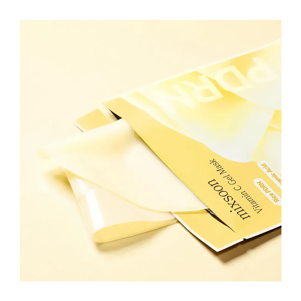 A close-up of a sheet mask package in pale yellow and white, with a translucent peel-off mask partially peeled back, on a matching light cream background.