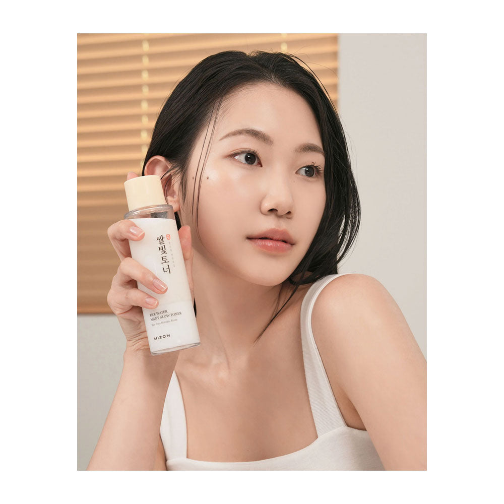 A woman in a white tank top holds a clear bottle of skincare serum with a beige cap in front of her face. The background features a soft beige wall and horizontal wooden blinds.