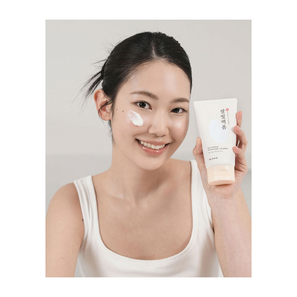 A woman with dark hair tied back wears a white tank top and holds up a cream-colored skincare tube while smiling. She has white cream smeared on her cheek.