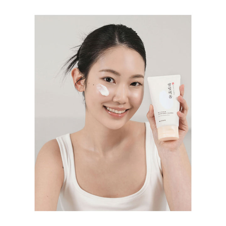 A woman with dark hair tied back wears a white tank top and holds up a cream-colored skincare tube while smiling. She has white cream smeared on her cheek.
