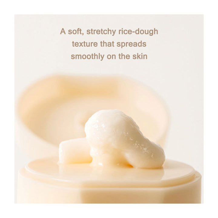 Creamy off-white cosmetic texture dollop on a beige jar lid with soft-focus background and text describing its smooth, stretchy rice-dough consistency.