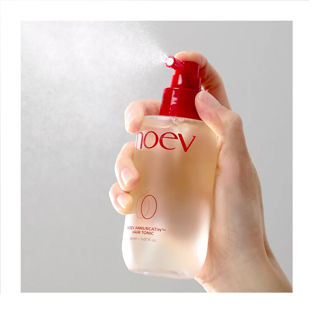 A hand holding a frosted bottle of Noev 10% Ammoniacalrin Hair Tonic, spraying a fine mist against a light gray background.