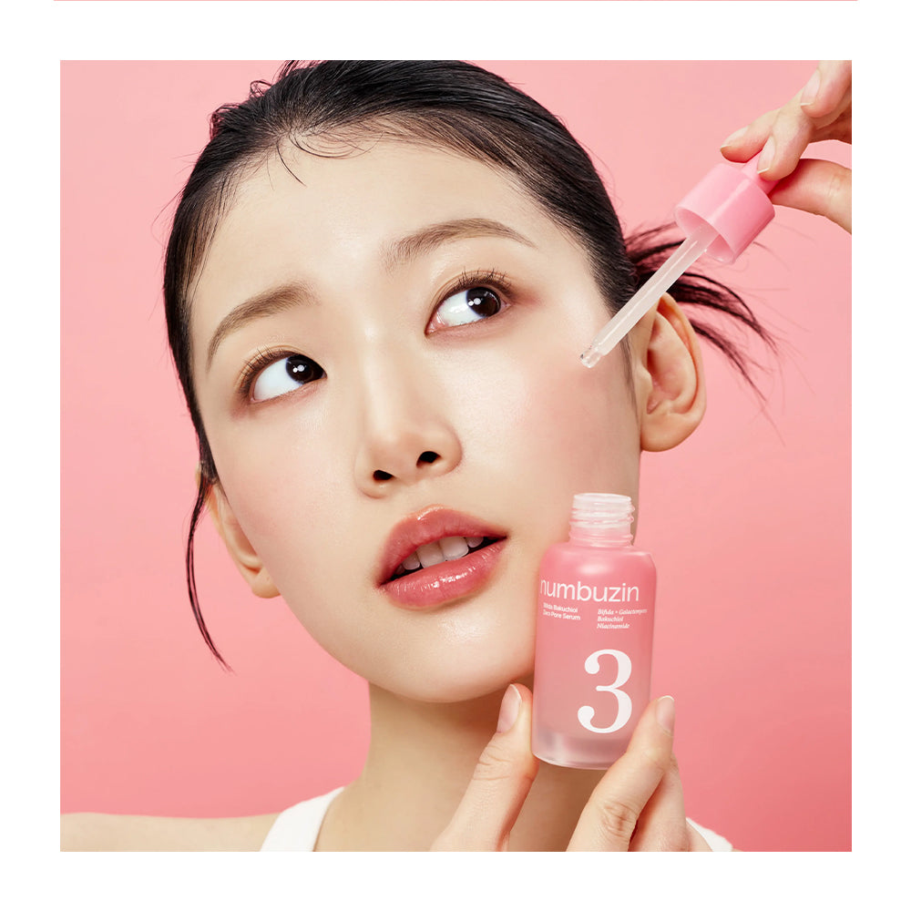 A close-up beauty shot of a woman with dark hair holding a pink bottle labeled 'umbuzin 3' while another hand applies a clear liquid from a dropper to her cheek against a solid pink background.