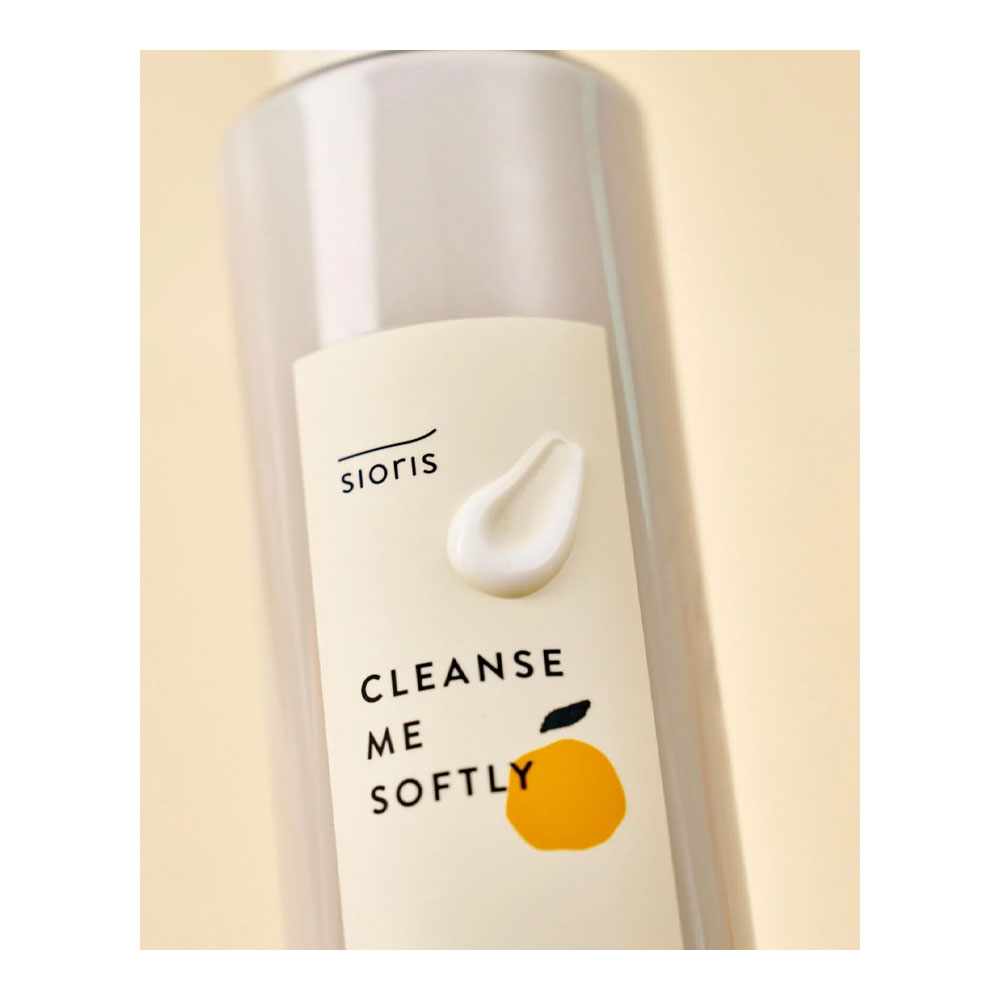 A close-up of a Sioris Cleanse Me Softly cleansing bottle with a cream-colored label featuring a white droplet graphic and a yellow citrus illustration, set against a soft beige background.
