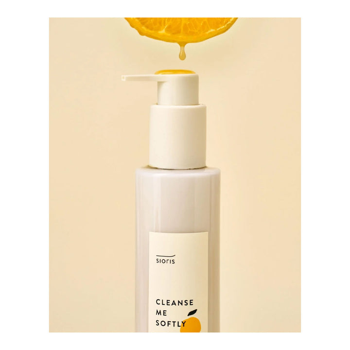 Sioris Cleanse Me Softly facial cleanser in a frosted white pump bottle, with a slice of orange dripping with juice above the pump, set against a warm beige background.