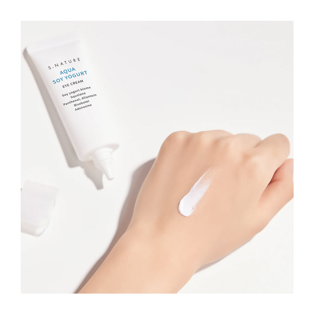 A white tube of S-Nature Aqua Soy Yogurt Eye Cream with its cap off, shown next to a hand with a dollop of the white cream on a clean white background.
