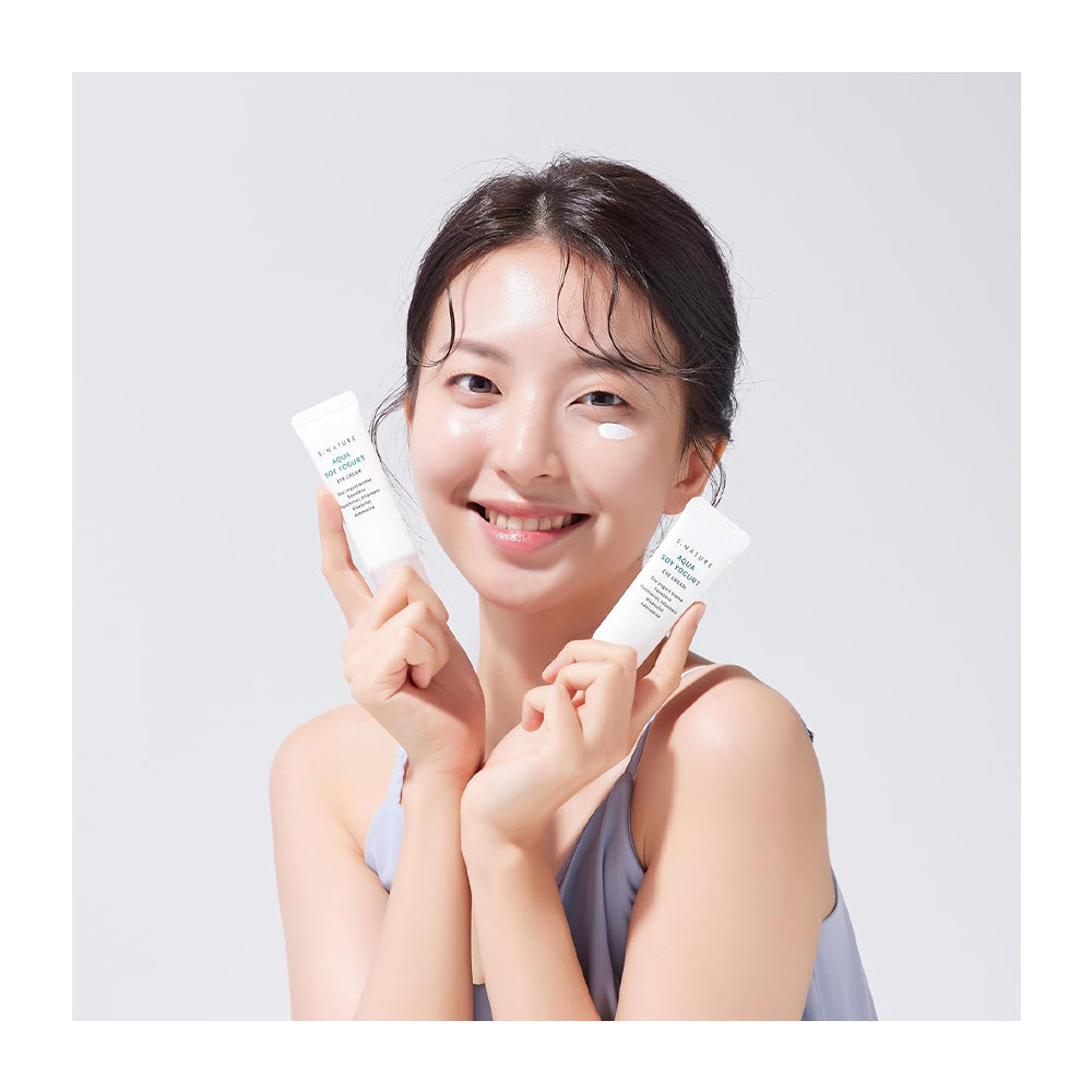A smiling woman with clear under-eye cream holds two white skincare tubes in both hands against a light gray background.