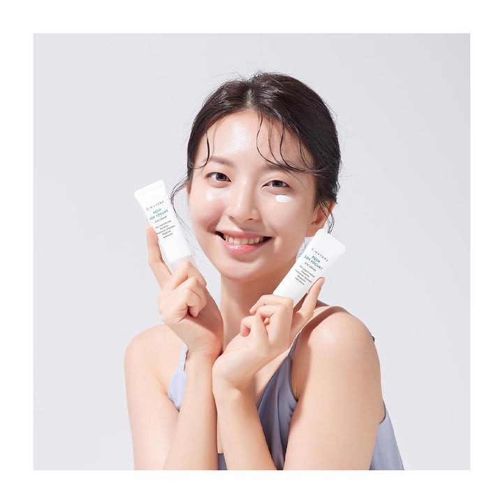 A smiling woman with clear under-eye cream holds two white skincare tubes in both hands against a light gray background.
