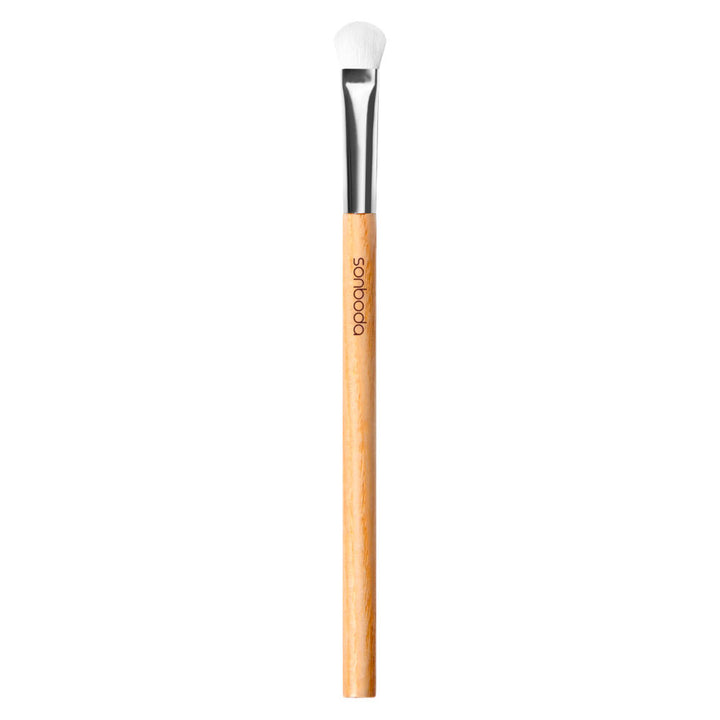 A single makeup brush with a light natural wood handle, a silver ferrule, and white bristles, shown vertically on a plain white background. The brand name &