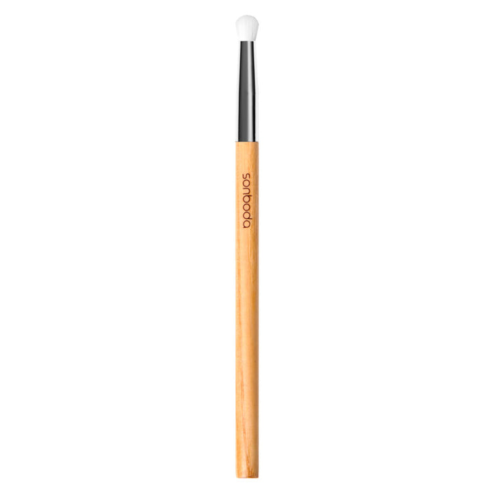 A single makeup brush with a light natural wood handle, a black ferrule, and a white bristle tip, shown vertically on a plain white background. The brand name &