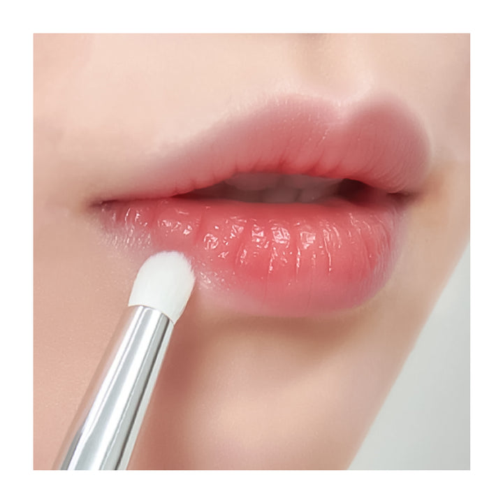 Close-up of glossy pink lips with a white-tipped applicator brush applying lip product, shown against a light neutral background.