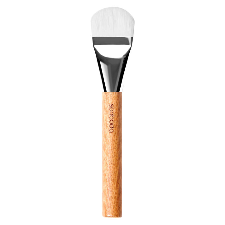 A flat makeup brush with white bristles, a shiny black ferrule, and a light brown wooden handle. The brand name &