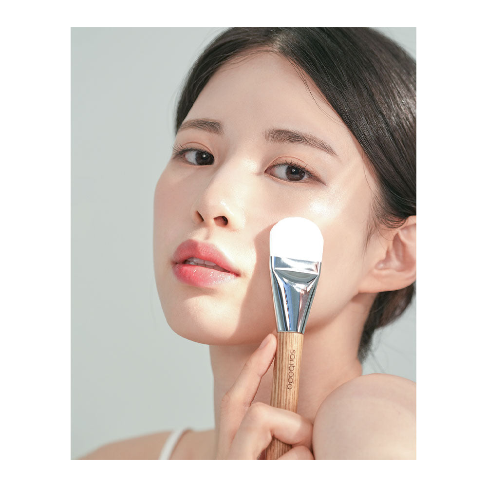 A close-up lifestyle shot of a woman with clear skin holding a makeup brush with a white silicone bristle head and a light wood handle against a soft gray background.