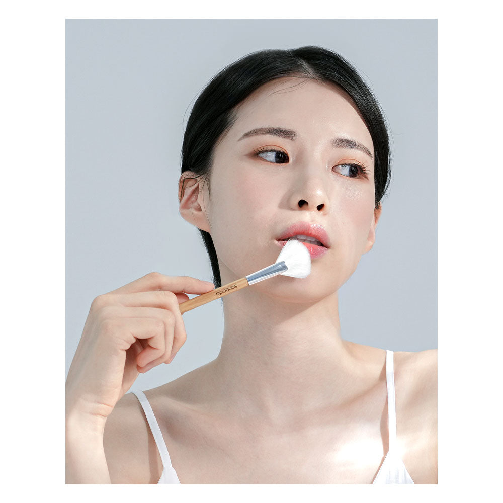 A woman with dark hair pulled back applies a white cream or powder from a makeup brush to her cheek, wearing a white spaghetti-strap top against a light gray background.