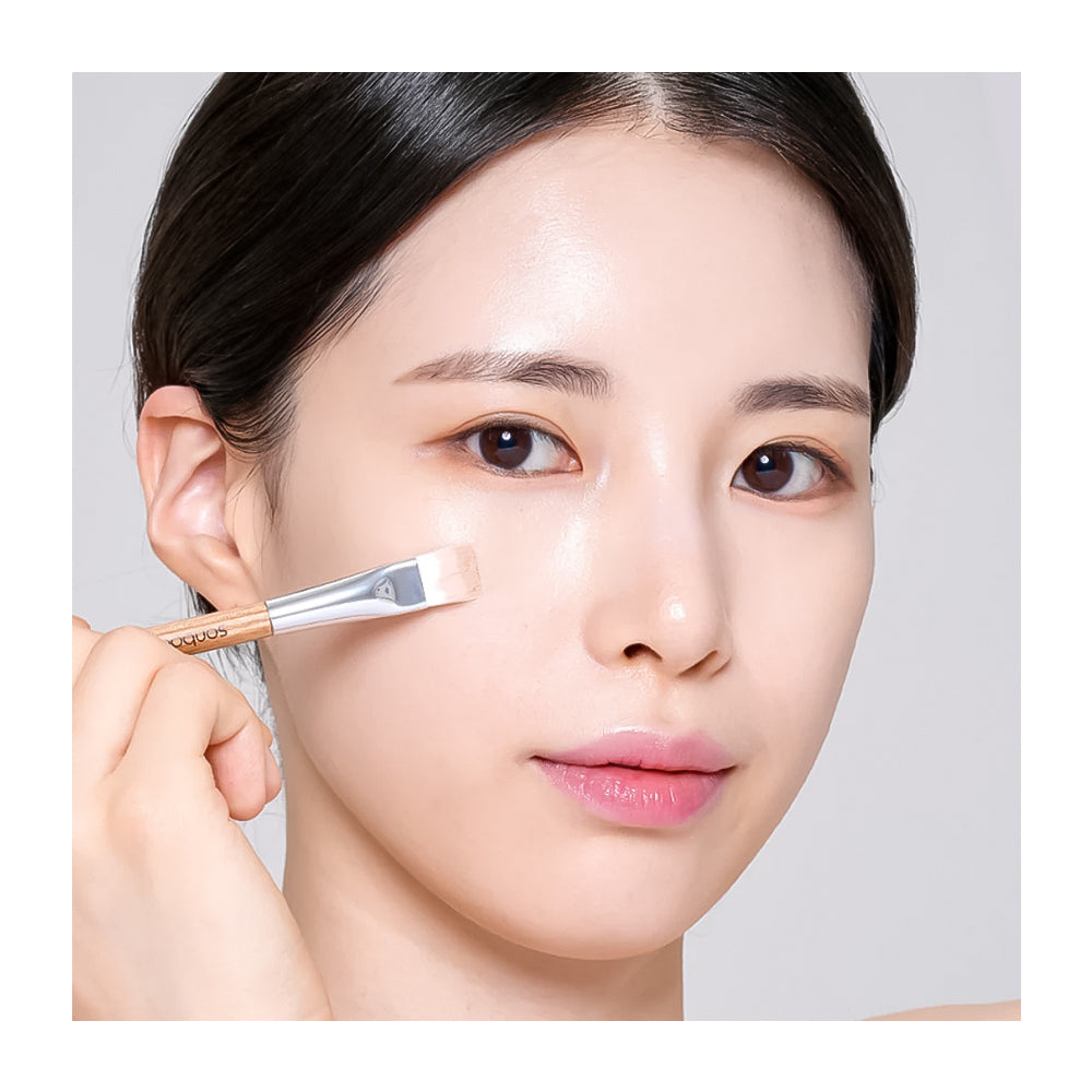 A close-up of a woman applying a light makeup product with a small angled brush to her cheek, set against a plain light gray background.