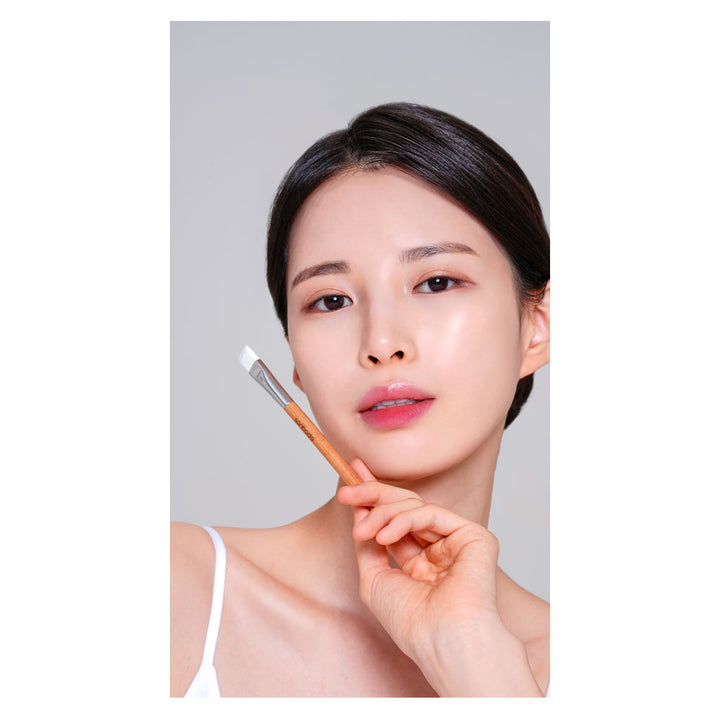 A close-up portrait of a woman with clean, radiant skin holding a small makeup brush near her chin, set against a plain light gray background.