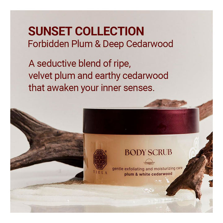 A jar of Tyla Body Scrub from the Sunset Collection, Forbidden Plum &amp; Deep Cedarwood blend, styled with natural wood branches on a reflective white surface against a light beige background with descriptive text.