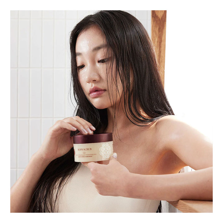 A woman with wet dark hair holds a cosmetic jar near her face in a bright bathroom setting with white tiled walls.