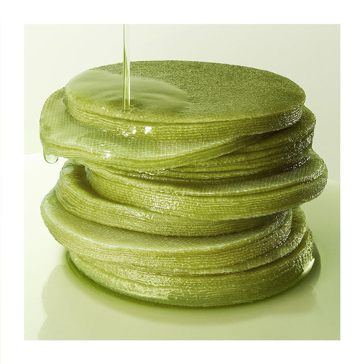 A stack of translucent green skincare patches or pads with a glossy, wet texture, with a clear liquid being poured onto the top piece against a pale green background.