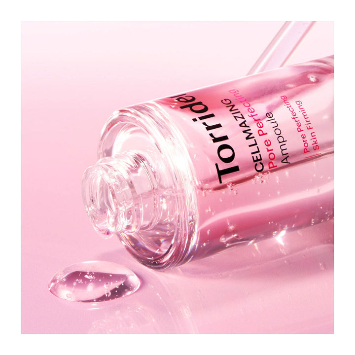 A clear glass bottle of Torrida Hyaluhydrating Perfecting Skin Serum lying on its side with pink serum dripping from the open cap onto a reflective pink surface.